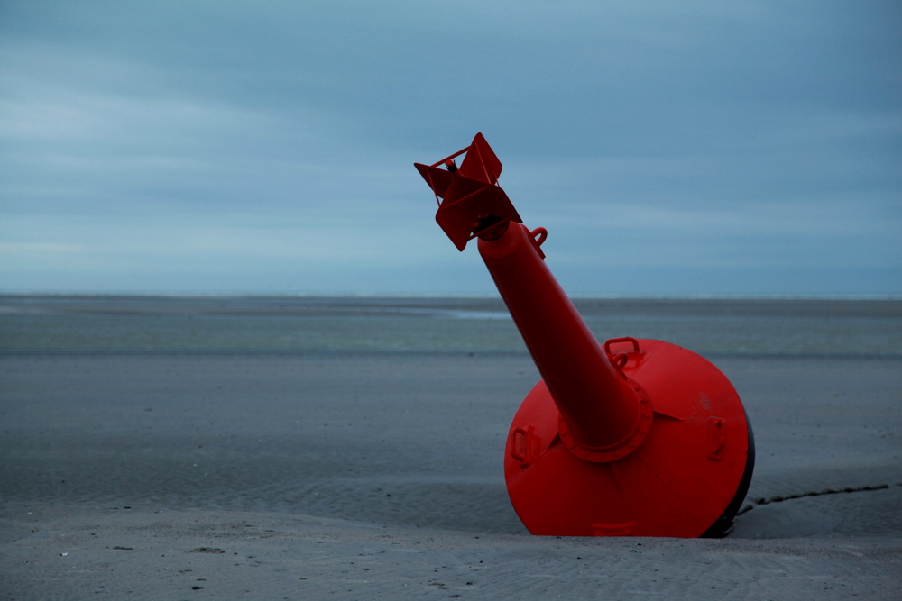 Posée sur le sable par marée basse, solidement arrimée au sol par une chaîne, la bouée de métal rouge indique le bord du chenal, utilisable par marée haute.

Le Touquet.